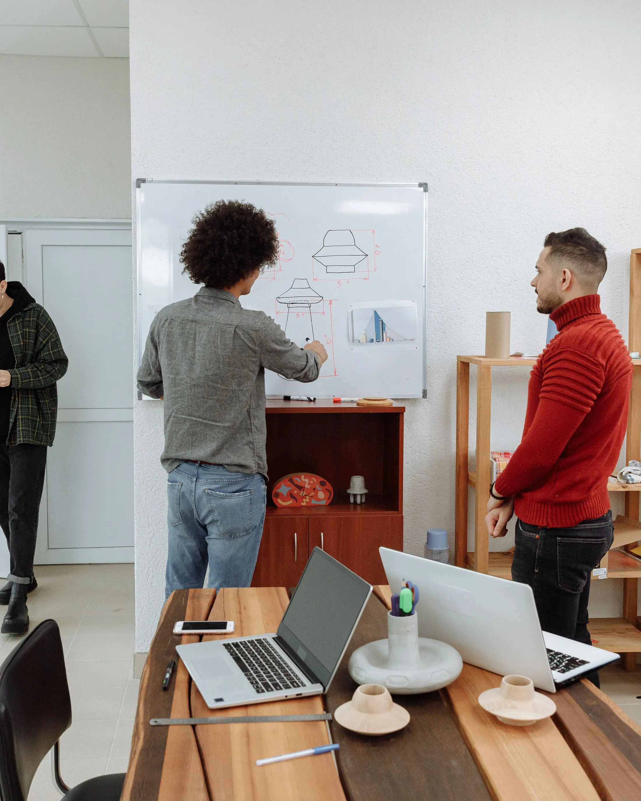 A developer sketching a diagram on a whiteboard while a colleague looks on, laptops on the desk in front